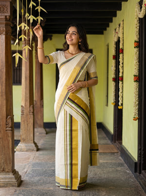 A woman wearing a traditional Indian saree stands in a corridor with wooden pillars. The saree is off-white/cream with a mustard yellow border and green stripes, paired with a matching off-white blouse.