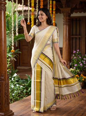 A woman modeling a cream-colored saree with yellow and purple stripes, standing in a traditional wooden porch decorated with marigold garlands.