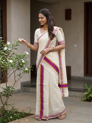 A woman wearing a cream-colored cotton saree with purple, pink, and gold borders, standing outdoors in front of a building with stone walls and a small flowering plant in the foreground.