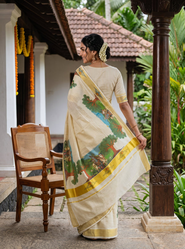 A woman wearing a cream-colored saree with a green and blue landscape print and a yellow border, paired with a beige blouse, standing on a traditional porch.