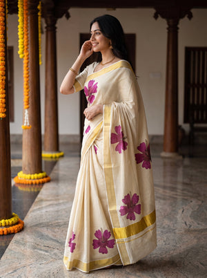 A woman wearing a cream-colored silk saree with purple floral prints and gold borders, standing in a traditional courtyard with wooden pillars and yellow garlands.