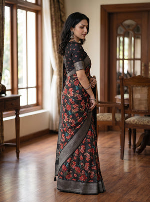 A woman wearing a dark floral saree with a silver border, standing in a traditional indoor room with wooden floors and windows.