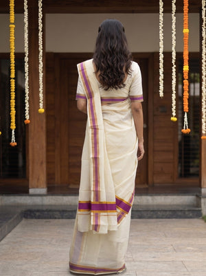 A woman viewed from behind wearing a cream-colored cotton saree with purple and gold border stripes, standing in front of a traditional wooden doorway decorated with hanging marigold garlands.