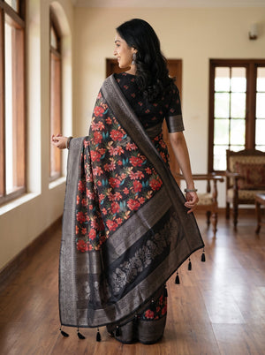 A woman wearing a black floral saree with red and orange flowers, green leaves, and a wide metallic silver border, standing in a bright room with wooden floors and windows.