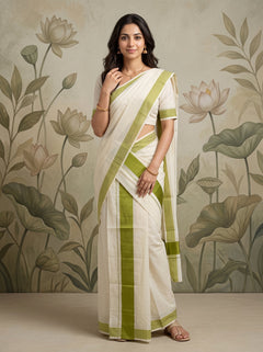 A woman modeling a white and olive green saree with a matching short-sleeve blouse, standing against a beige wall with a floral lotus pattern.