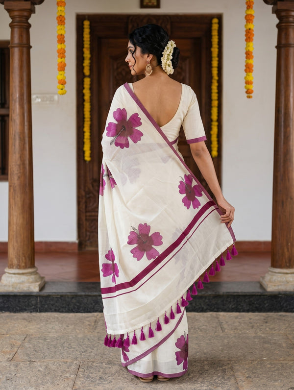A woman wearing a white and magenta floral saree, photographed from behind in front of a traditional doorway with wooden pillars and yellow marigold garlands.