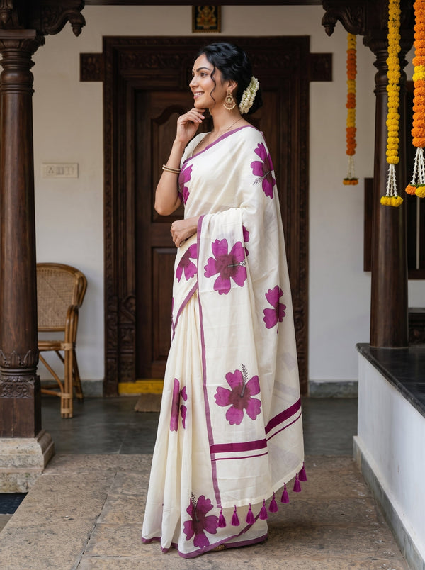 A woman wearing a cream-colored cotton saree with large magenta floral prints and border details, standing in a traditional courtyard with dark wooden pillars.