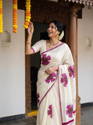 A woman wearing a white saree with large magenta floral prints and border details, standing in a traditional doorway and touching a hanging marigold garland.
