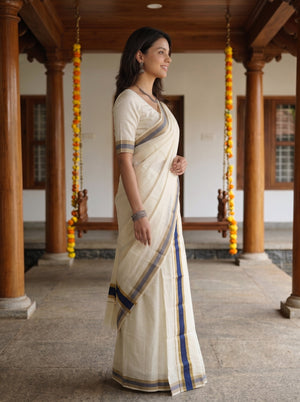 A woman wearing a cream-colored cotton saree with blue and gold stripes, standing in profile in a traditional courtyard with wooden pillars and hanging marigold garlands.