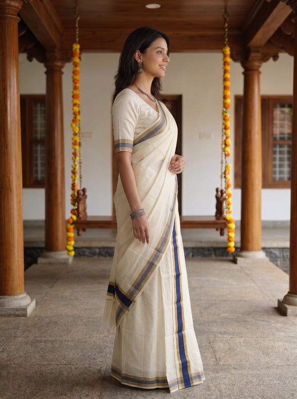 A woman wearing a cream-colored cotton saree with blue and gold stripes, standing in profile in a traditional courtyard with wooden pillars and hanging marigold garlands.