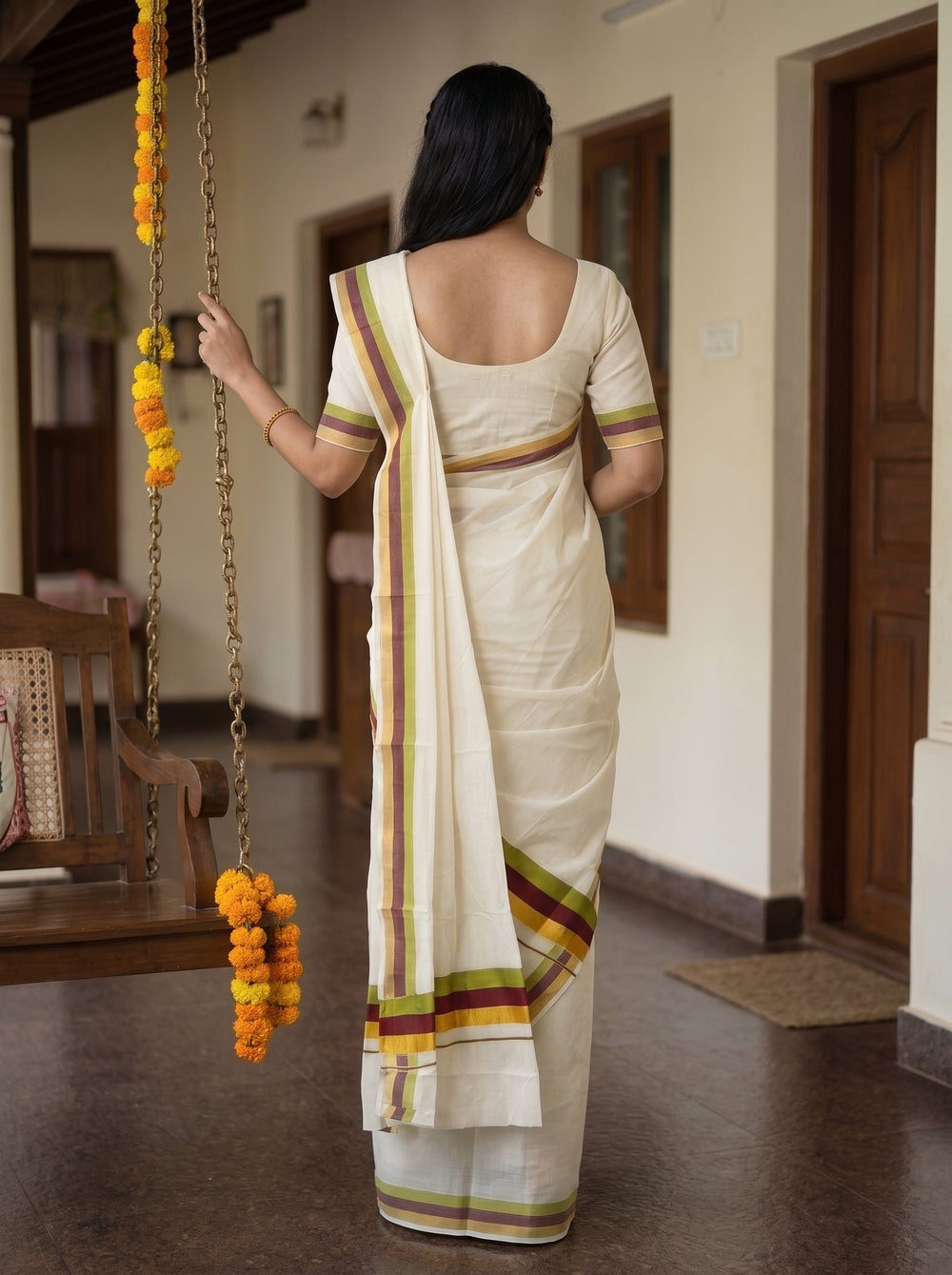 A woman wearing a cream-colored traditional Indian saree with green, yellow, and maroon stripes stands in a hallway holding a hanging flower garland.