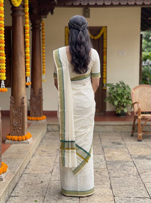 A woman viewed from behind wearing a cream-colored cotton saree with green and gold border accents, standing in a courtyard decorated with marigold garlands.