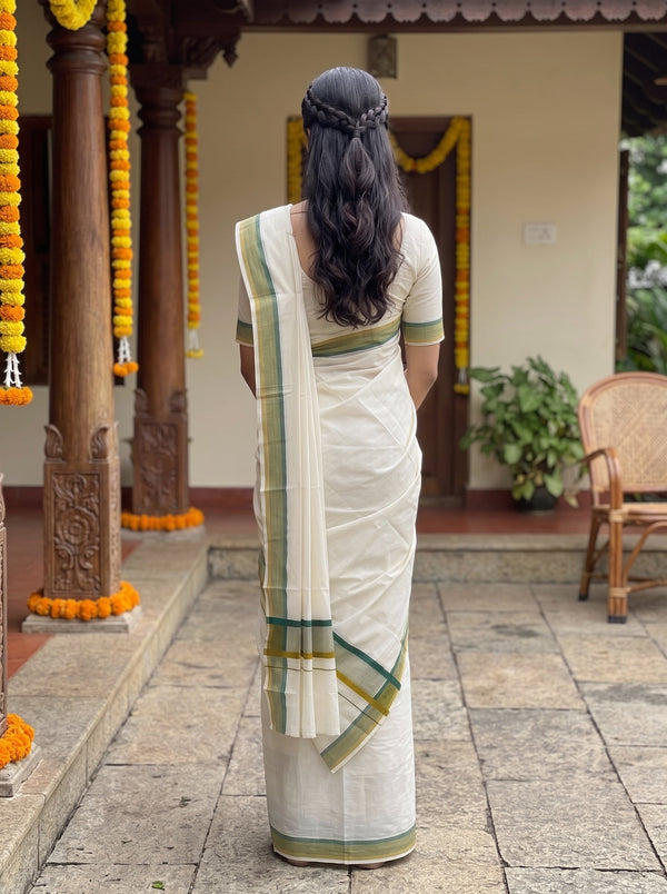 A woman viewed from behind wearing a cream-colored cotton saree with green and gold border accents, standing in a courtyard decorated with marigold garlands.