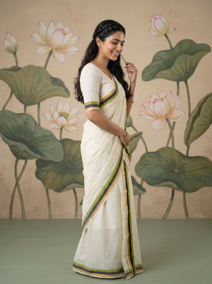A woman wearing a cream-colored cotton saree with green and yellow striped borders, standing in front of a beige wall decorated with painted lotus flowers and leaves.