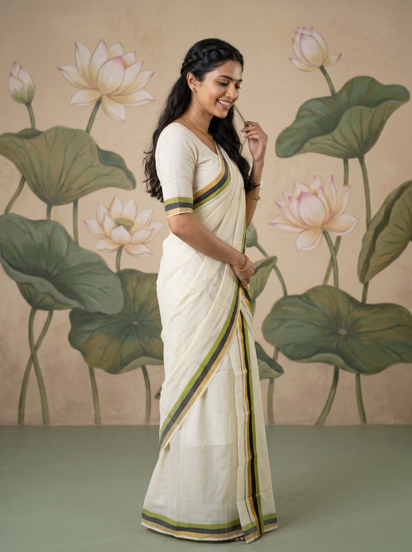 A woman wearing a cream-colored cotton saree with green and yellow striped borders, standing in front of a beige wall decorated with painted lotus flowers and leaves.