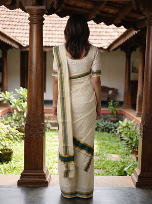 A woman viewed from behind wearing a cream-colored cotton saree with green and gold stripes, standing in a traditional courtyard framed by carved wooden pillars.