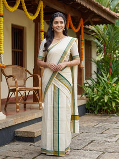 A woman wearing a cream-colored cotton saree with green and gold stripes, standing in front of a traditional house courtyard decorated with yellow and orange marigold garlands.