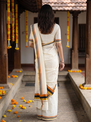 A woman viewed from behind wearing a cream-colored cotton saree with orange, yellow, and black striped borders, standing in a traditional courtyard decorated with marigold garlands.
