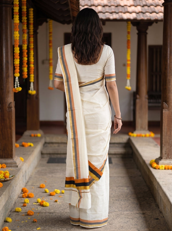 A woman viewed from behind wearing a cream-colored cotton saree with orange, yellow, and black striped borders, standing in a traditional courtyard decorated with marigold garlands.