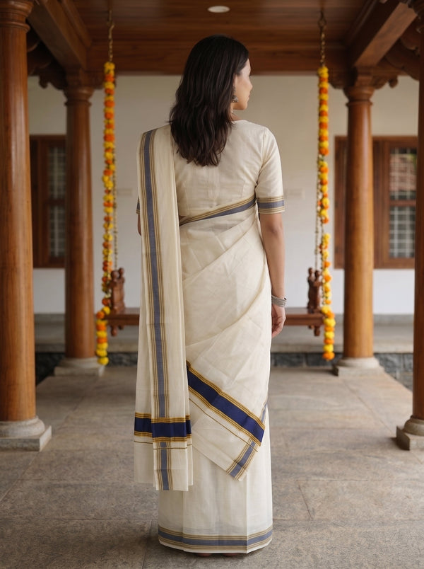 A woman viewed from behind wearing a cream-colored cotton saree with blue and gold stripes, standing in a traditional courtyard with wooden pillars and hanging marigold garlands.