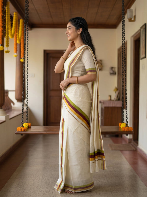 A woman wearing a cream-colored cotton saree with green, yellow, and orange border stripes stands in profile in a traditional Indian indoor hallway with hanging swings.