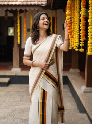 A woman wearing a cream-colored cotton saree with orange, green, and black stripes, standing in a courtyard decorated with yellow marigold garlands.