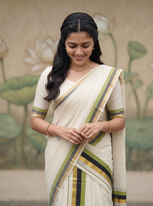 A woman wearing a cream-colored cotton saree with olive green, black, and mustard yellow stripes, standing in front of a painted lotus wall.