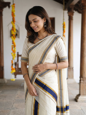 A woman wearing a cream-colored cotton saree with blue and gold stripes, standing in a traditional courtyard with wooden pillars and hanging marigold garlands.