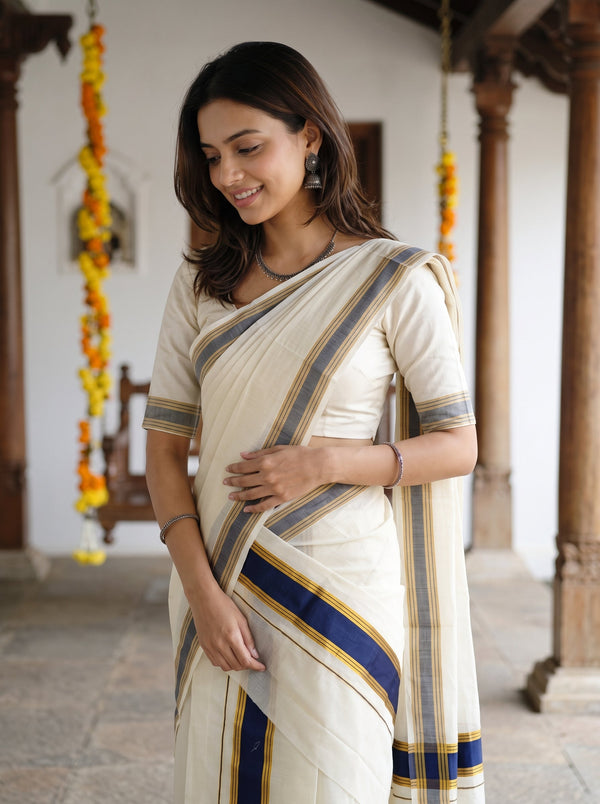 A woman wearing a cream-colored cotton saree with blue and gold stripes, standing in a traditional courtyard with wooden pillars and hanging marigold garlands.