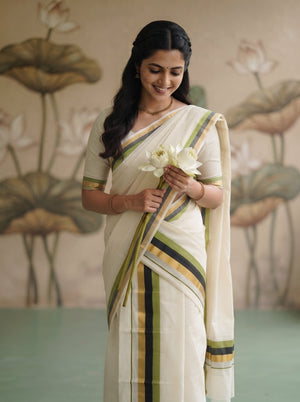 A woman wearing a cream-colored cotton saree with green and gold stripes, holding white lotus flowers against a soft-focus lotus wallpaper background.