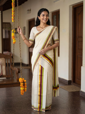 A woman wearing a cream-colored traditional Indian saree with green, yellow, and maroon stripes stands in a hallway holding a hanging flower garland.