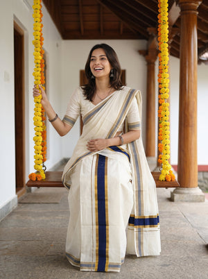 A woman wearing a cream-colored cotton saree with navy blue and gold stripes, standing on a traditional wooden swing decorated with yellow flower garlands.