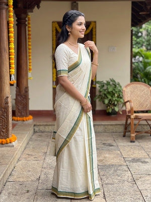 A woman wearing a cream-colored cotton saree with green and gold borders, standing in a traditional courtyard setting.