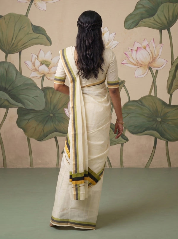 A woman viewed from behind wearing a cream-colored saree with green and yellow striped borders, standing in front of a wall mural featuring large green lotus leaves and pink-white lotus flowers.