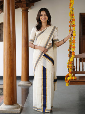 A woman wearing a cream-colored cotton saree with navy blue and gold borders, standing in a traditional indoor setting with wooden pillars and holding a yellow and orange marigold garland.