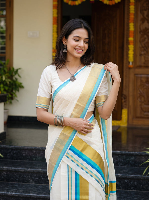A woman wearing a cream-colored sari with blue and gold stripes, standing on black marble steps in front of a decorated wooden doorway.
