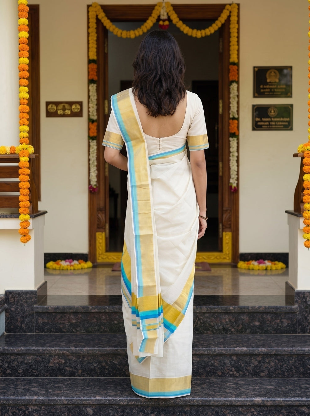 A woman wearing a cream-colored cotton saree with blue, yellow, and white stripes stands on stone steps in front of a doorway decorated with marigold garlands.