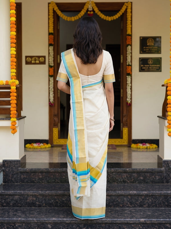 A woman viewed from behind wearing a white saree with a yellow and blue border, standing on dark stone steps in front of a decorated doorway with marigold garlands.