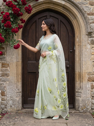 A woman wearing a light sage green embroidered saree stands in front of a rustic stone wall and dark wooden door, with red flowers hanging on the left.