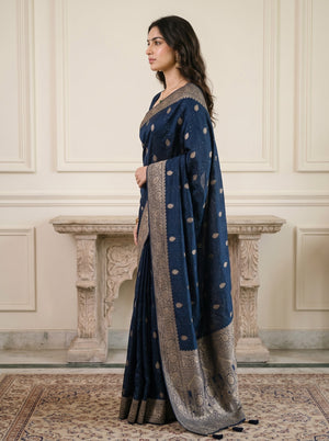 A woman wearing a dark blue traditional sari with gold and silver motifs and a wide metallic border, standing in profile in front of a cream-colored paneled wall and stone mantel.