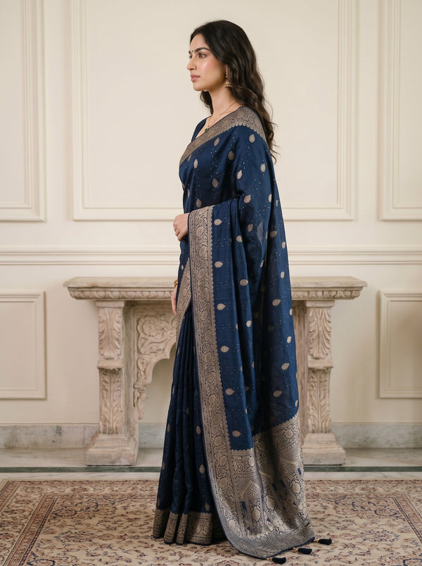 A woman wearing a dark blue traditional sari with gold and silver motifs and a wide metallic border, standing in profile in front of a cream-colored paneled wall and stone mantel.