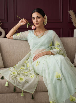 A woman seated on a beige sofa wearing a pale mint green embroidered saree with floral motifs and tassel details, holding a pink rose against a dark burgundy paneled wall background.