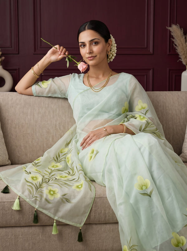 A woman seated on a beige sofa wearing a pale mint green embroidered saree with floral motifs and tassel details, holding a pink rose against a dark burgundy paneled wall background.