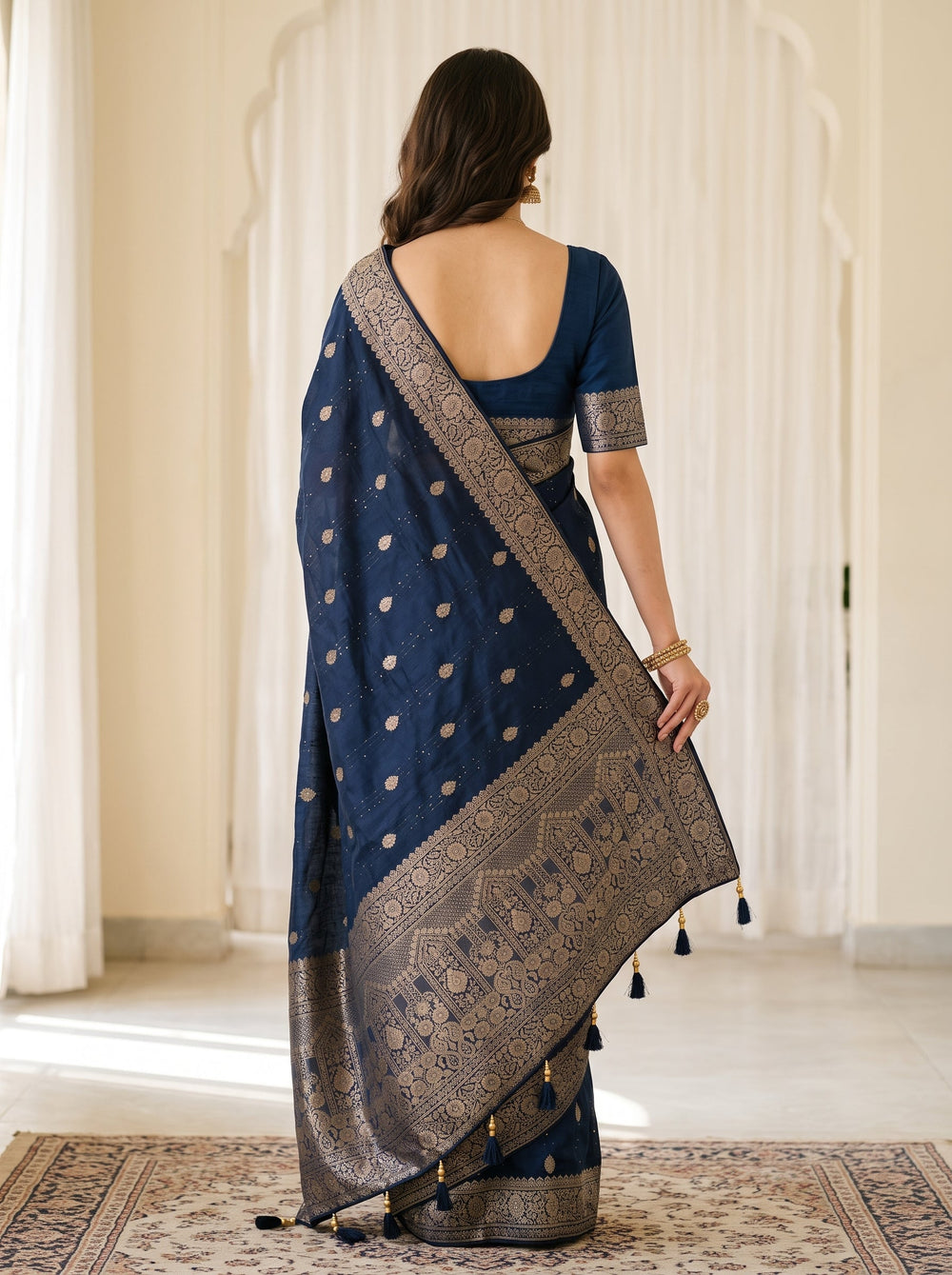 A woman modeling a navy blue saree with silver metallic border and motifs, standing in front of a cream-colored paneled wall and stone mantel.
