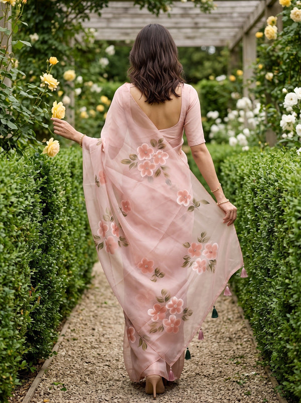A woman viewed from behind wearing a light pink saree with floral embroidery, walking down a gravel path lined with green hedges and yellow roses in a garden.