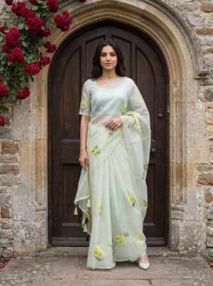 A woman stands in front of a stone archway and dark wooden door wearing a pale mint green saree with yellow floral prints. Red flowers and green leaves hang on the upper left side of the frame.