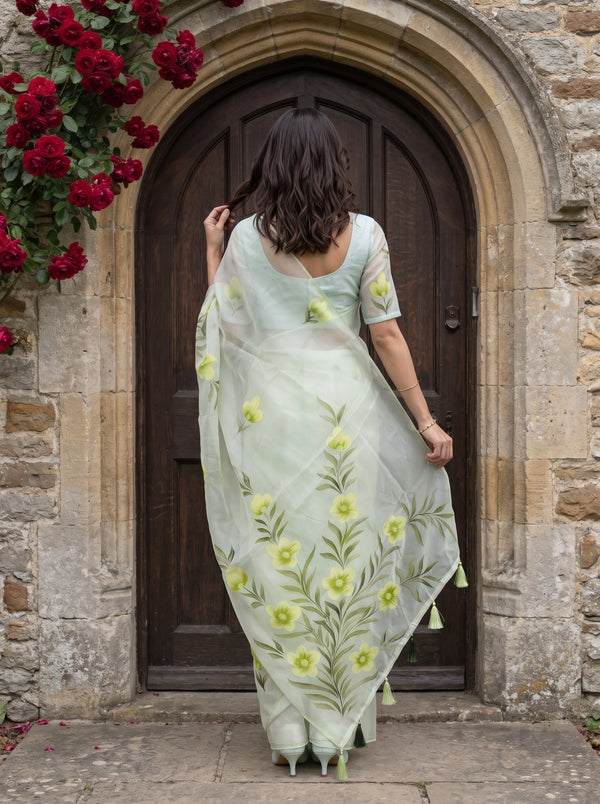 A woman viewed from behind wearing a pale sage green organza saree with yellow floral embroidery, standing in front of a rustic stone archway and dark wooden door.