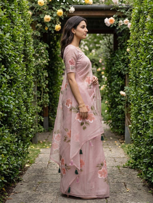 A woman wearing a dusty pink floral printed saree stands in profile on a stone path surrounded by a lush green garden with rose arches.
