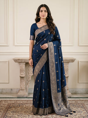 A woman modeling a navy blue saree with silver metallic border and motifs, standing in front of a cream-colored paneled wall and stone mantel.
