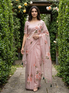A woman stands on a stone path between tall green hedges wearing a light pink floral saree with a matching blouse. The saree has pink and peach flowers with green leaves and dark green tassel ties.